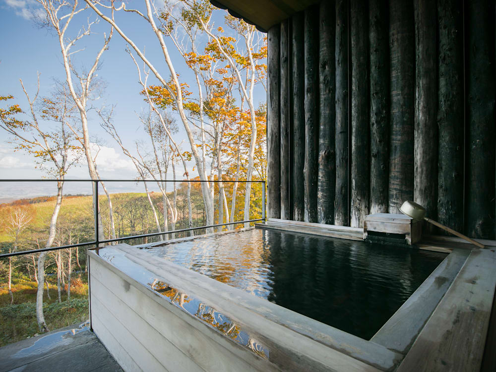 A serene outdoor Japanese onsen bath surrounded by nature, with steam rising gently from the water and a view of trees and misty hills in the background, creating a peaceful and tranquil atmosphere.
