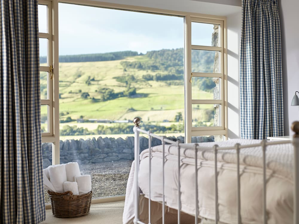 A bedroom with a white iron frame and white bedding. The view outside of rolling hills in Scotland