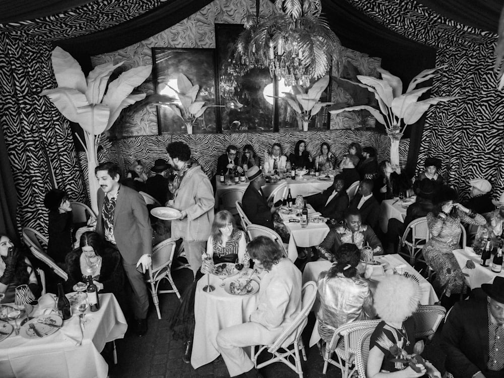 A black and white photo of diners in a restaurant. The walls are draped with leopard print material and the guests are dressed in glamorous Texan attire.