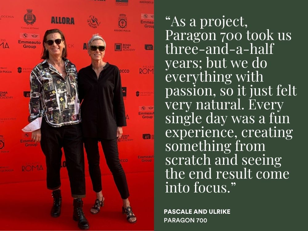Portrait of two women in dark shades, dark trousers and shirts, stood on a red carpet in front of a sponsor wall.