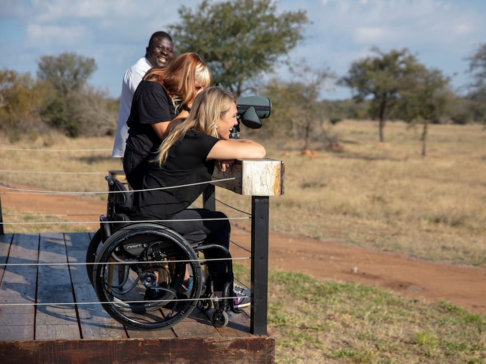 A man in a white top and two women in black clothing are on a wooden safari viewing point. The middle woman is using binoculars and the nearest woman is in a wheelchair. 