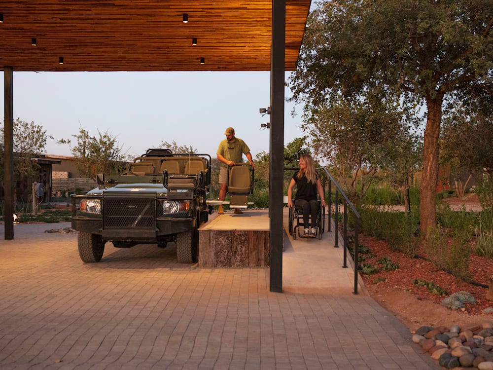 A woman in a wheelchair descends a purpose built ramp, next to a safari vehicle with accessible seating. The sun is setting and they are beneath a wooden covering.