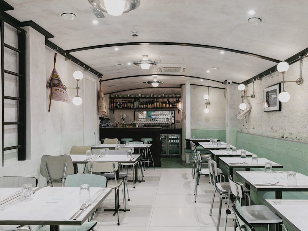 The inside of a restaurant, with white floors, ceilings, walls and tables, alongside mint green chairs and tiles running on the bottom half of the walls. A bar is in the back of the room.