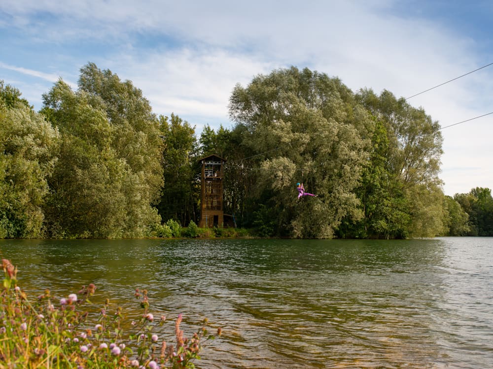 A child dressed in pink is riding a zip wire on a lake with trees in the background.