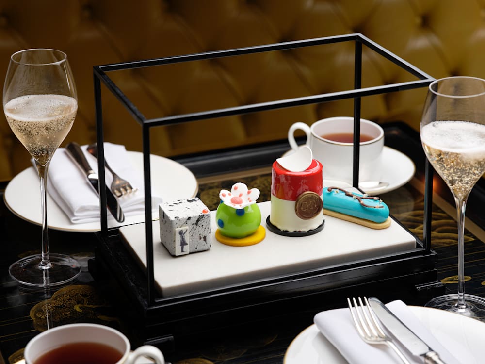 There are four small cakes on a plate, each replica of a different famous work of art, and around them are two glasses of wine and a cup of tea.  There is a yellow sofa behind the black table with the cake on it.