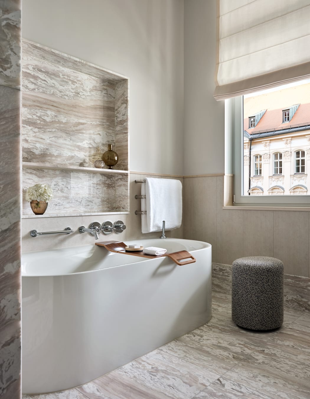 A white bathtub in a marble bathroom, with a window overlooking local area.