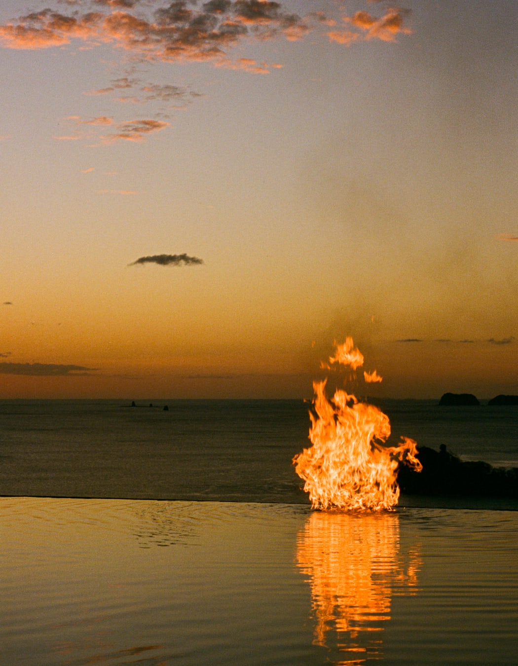 A fire burns by the edge of an infinity pool, looking over the ocean as the sun sets.