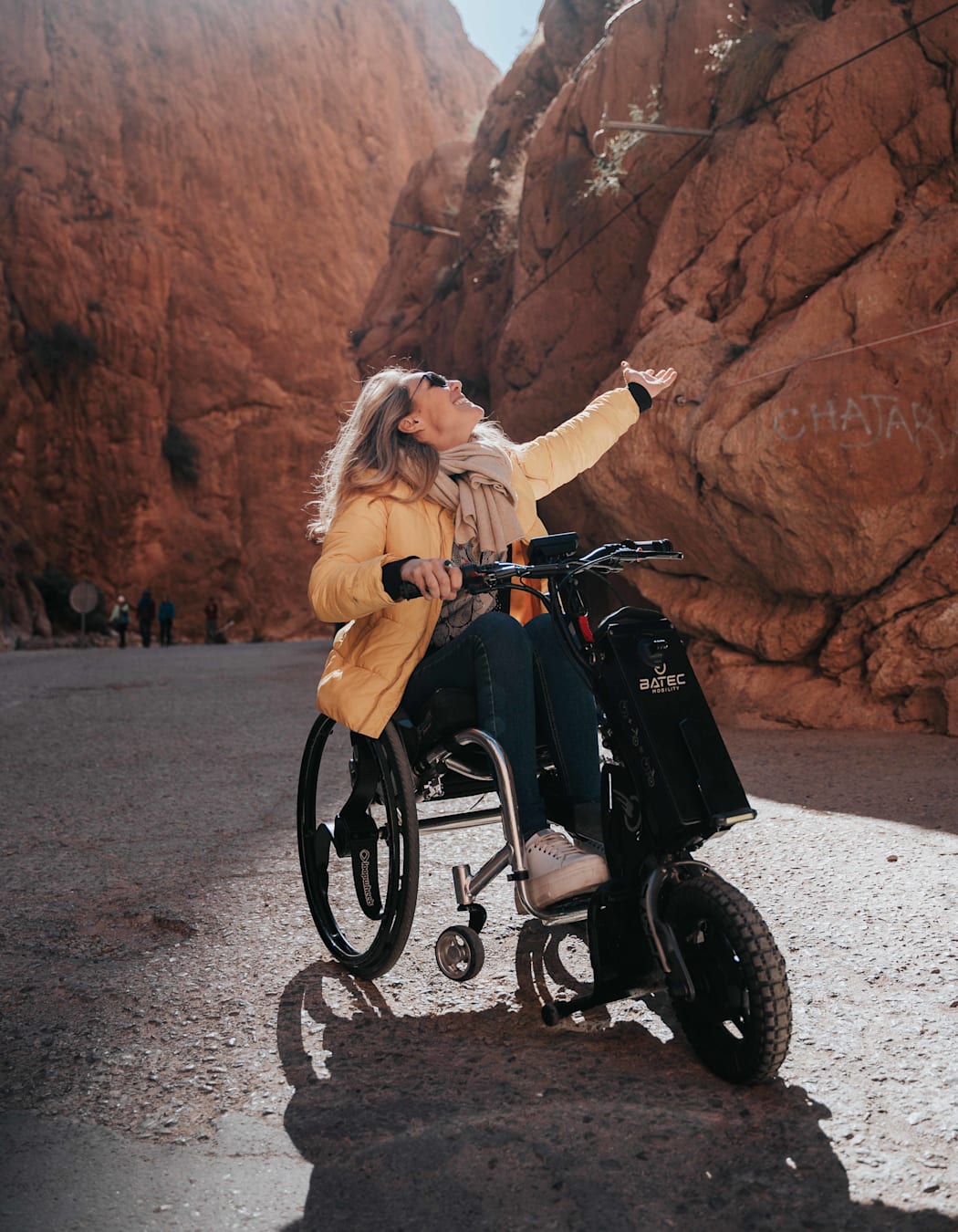 A happy woman in a yellow puffer jacket, blue jeans and white trainers sits on a three-wheeled mobility vehicle whilst gesturing upwards to the rocks in the ravine she is in. 