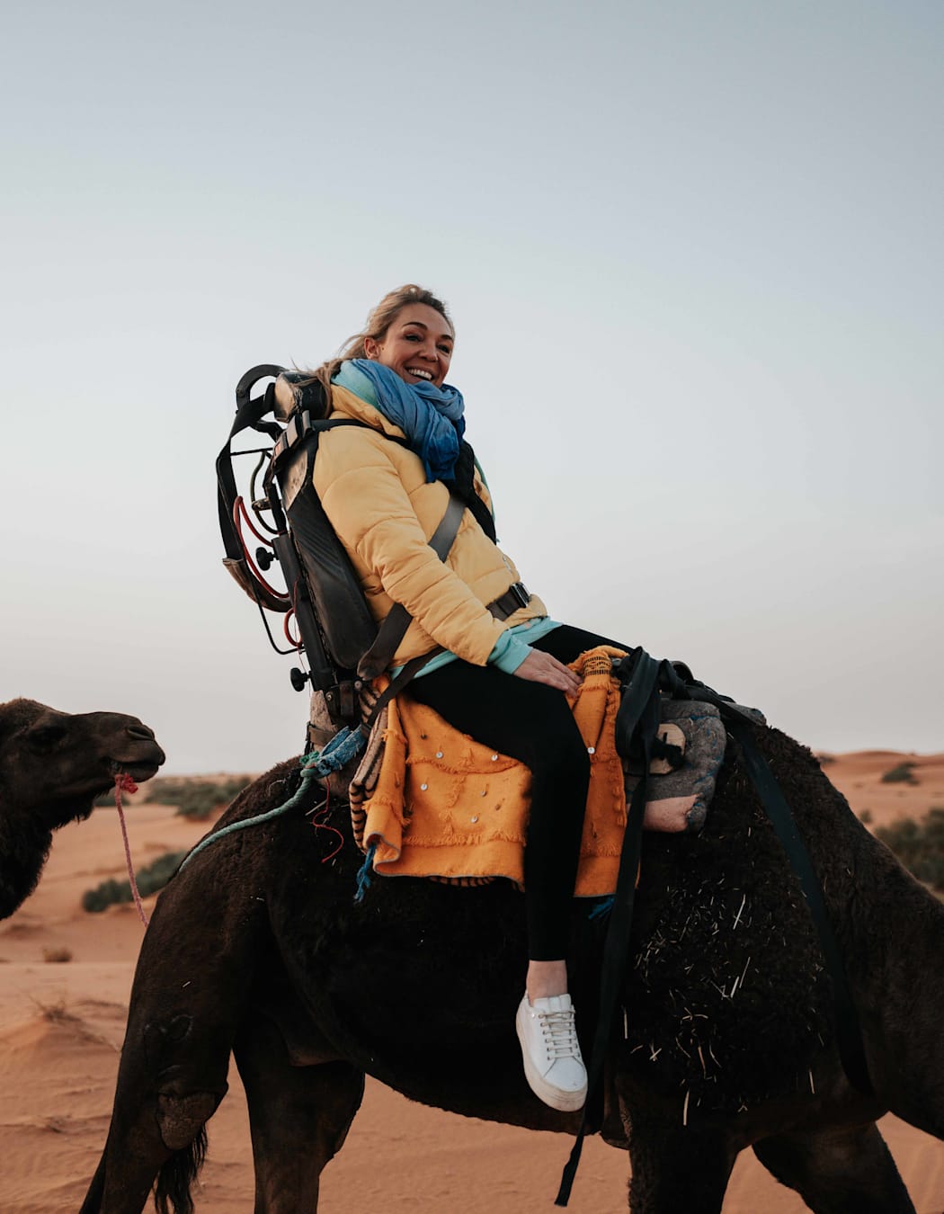 A woman in a yellow puffer jacket and blue scarf smiles as she rides a camel in the desert.