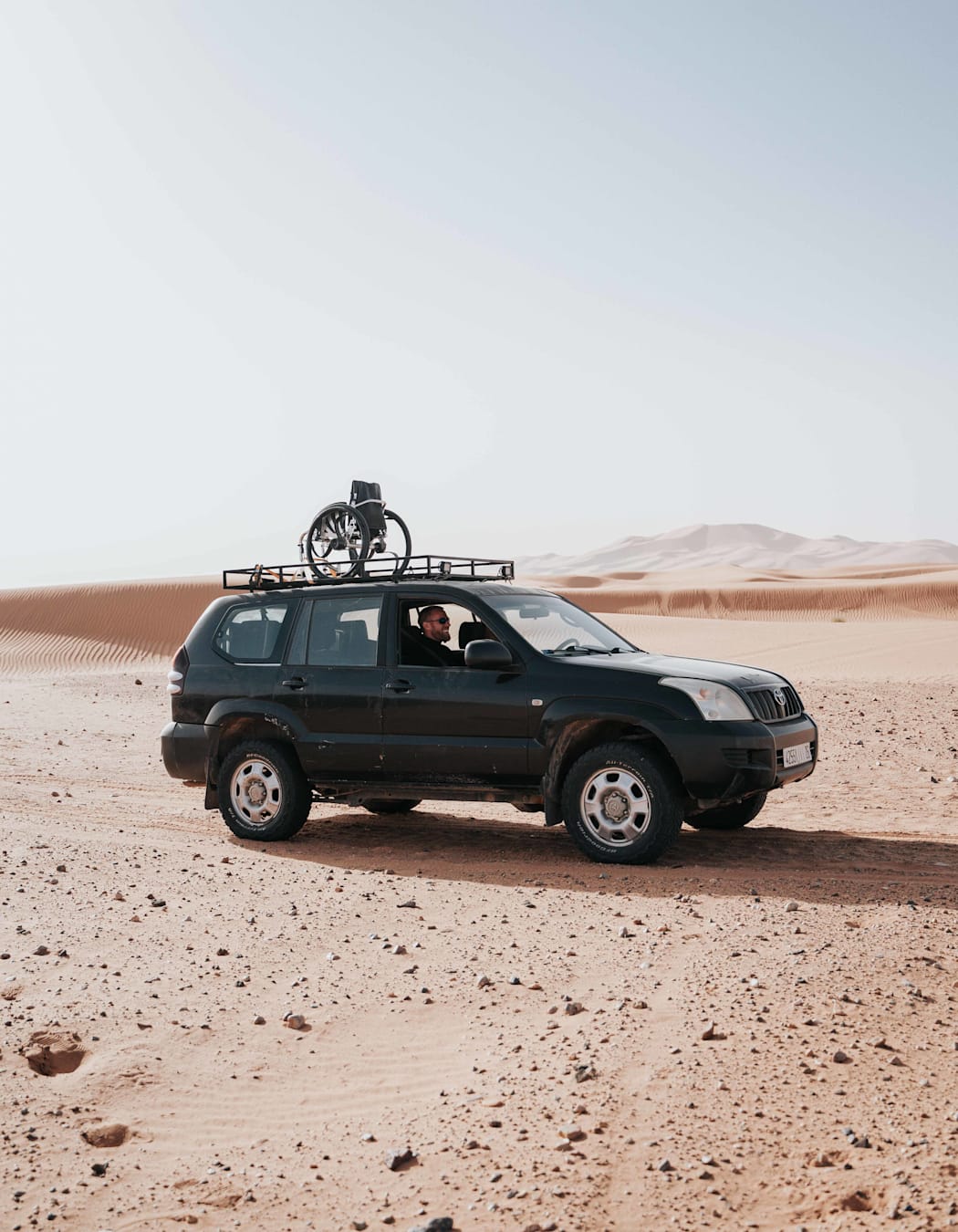 A side-profile photo of a jeep in the desert, carrying a wheelchair on it's roof.
