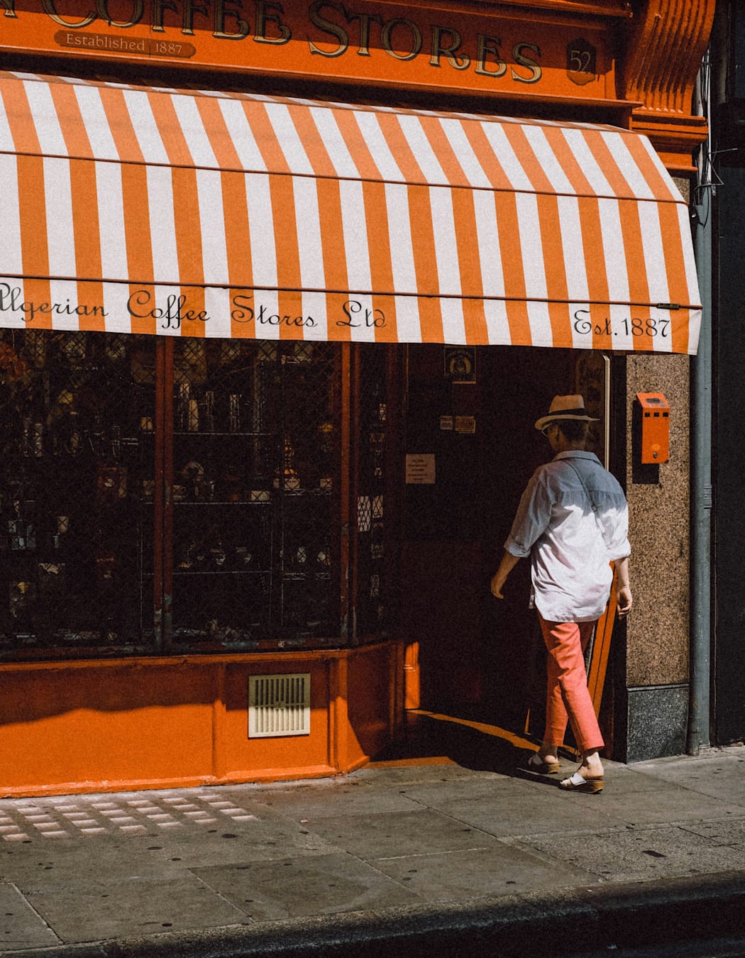 A woman wearing salmon coloured trousers and a white shirt enters into an orange coloured shop. The shop has a striped awning on the outside with the words 